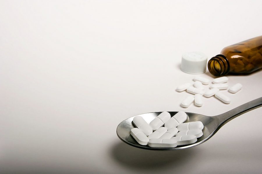 A bottle falls next to some pills in the background with a spoon in the foreground holding more pills.